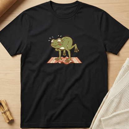 A black short-sleeve crewneck t-shirt laid flat on a light wood surface, featuring a cartoon graphic of a green frog standing on a red patterned rug. Minimalist yoga apparel by pemuyo.