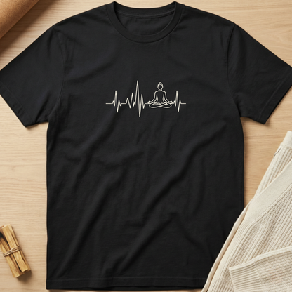 A black short-sleeve t-shirt laid flat on a light wood surface, featuring a white line-art graphic of a meditating figure integrated into an EKG heartbeat line across the chest. Minimalist yoga apparel by pemuyo.