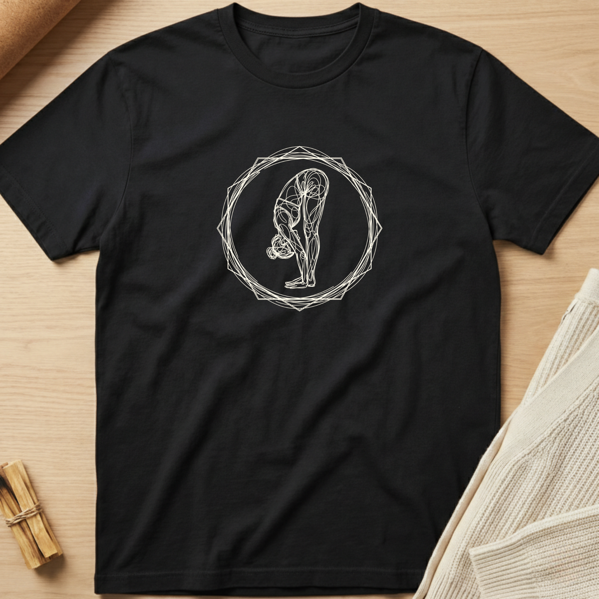 A black short-sleeve crewneck t-shirt with a white line art graphic of a person in a yoga pose, laid flat on a light wood background with neutral props. Minimalist yoga apparel by pemuyo.