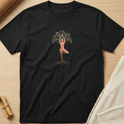 A black short-sleeve crewneck t-shirt laid flat on a light wood background, featuring a central graphic of a tree with green leaves and roots, with a pink figure posed inside the branches. Minimalist yoga apparel by pemuyo.