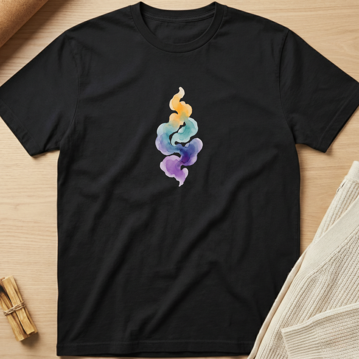 A black short-sleeve crewneck t-shirt laid flat on a light wood surface, featuring a colorful watercolor-style smoke graphic in yellow, blue, and purple on the center chest. Minimalist yoga apparel by pemuyo.