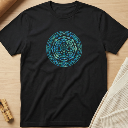 A black short-sleeve crewneck t-shirt laid flat on a light wood surface, featuring a circular mandala-style graphic in shades of blue and teal on the chest. Styled flat-lay props include a small bundle of wooden sticks in the bottom-left corner and a folded cream knit fabric in the bottom-right. Minimalist yoga apparel by pemuyo.