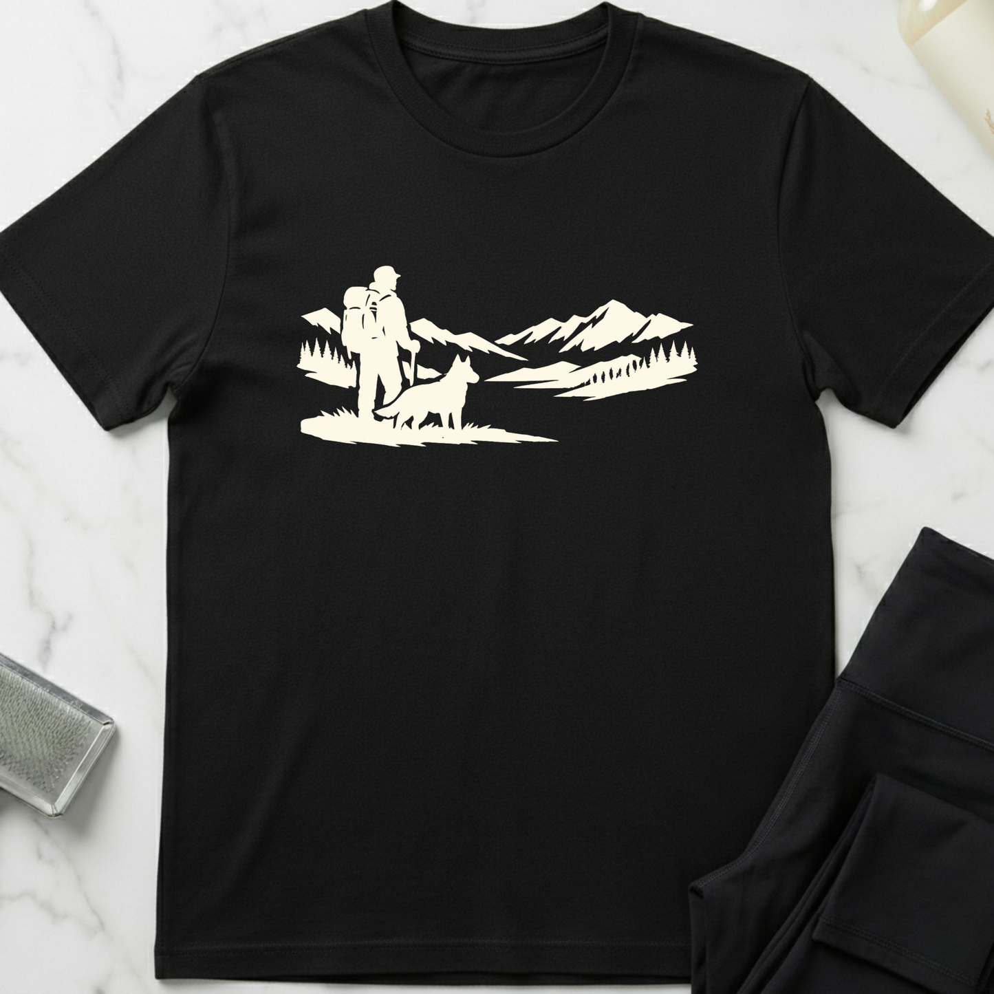 Black t-shirt with white graphic of a person and dog in a mountainous landscape on a marble surface. Minimalist dog apparel by pemuyo.