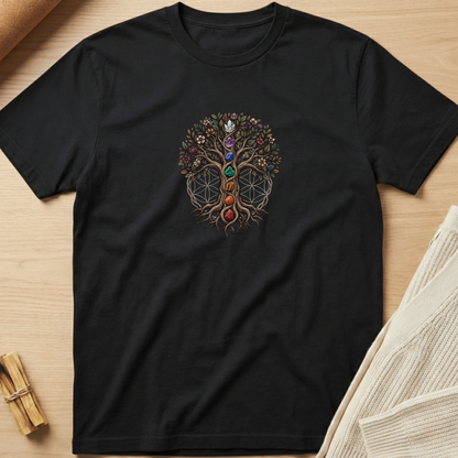 A black short-sleeve crewneck t-shirt laid flat on a light wood surface, featuring a colorful Tree of Life graphic with chakra symbols on the chest. Styled flat-lay props include a rolled beige fabric in the top-left and a folded cream knit blanket in the bottom-right. Minimalist yoga apparel by pemuyo.