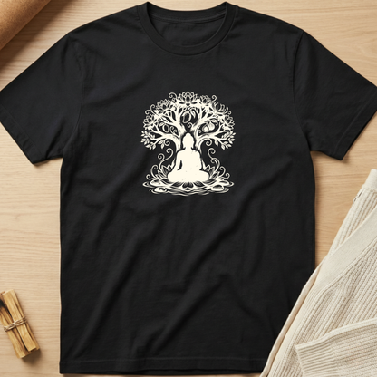 A black short-sleeve crewneck t-shirt with a white line-art graphic of a tree of life with a meditating figure, laid flat on a light wood background with styled props. Minimalist yoga apparel by pemuyo.