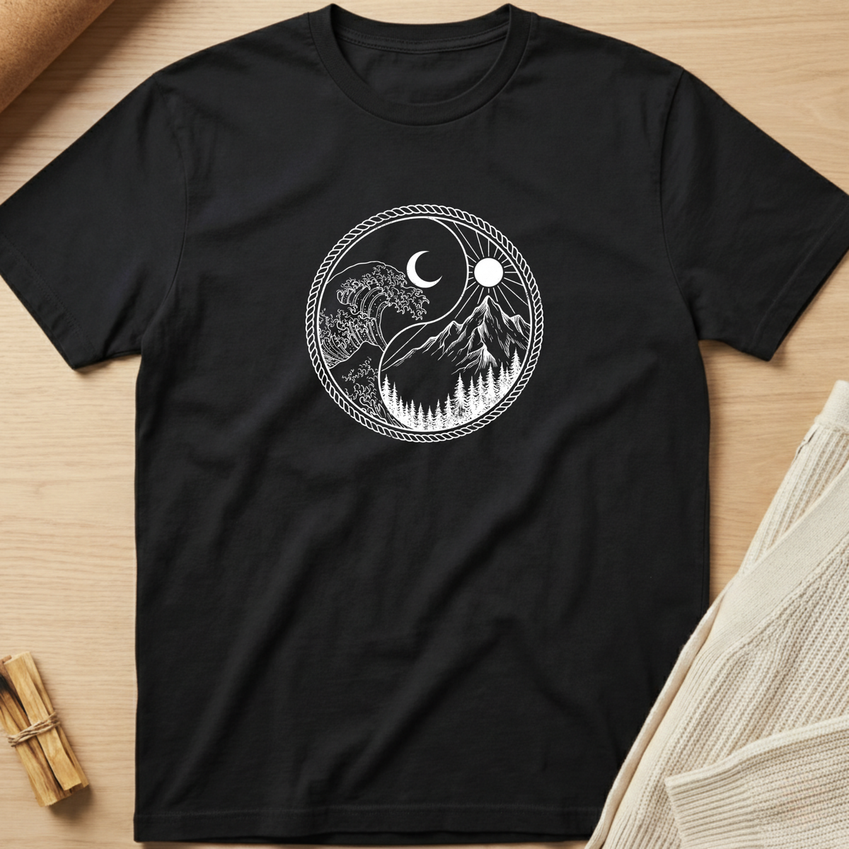 A black short-sleeve crewneck t-shirt laid flat on a light wood surface, featuring a white circular graphic of a yin-yang style design with a crescent moon, sun, mountain peaks, and pine trees. Minimalist yoga apparel by pemuyo.