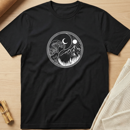 A black short-sleeve crewneck t-shirt laid flat on a light wood surface, featuring a white circular graphic of a yin-yang style design with a crescent moon, sun, mountain peaks, and pine trees. Minimalist yoga apparel by pemuyo.