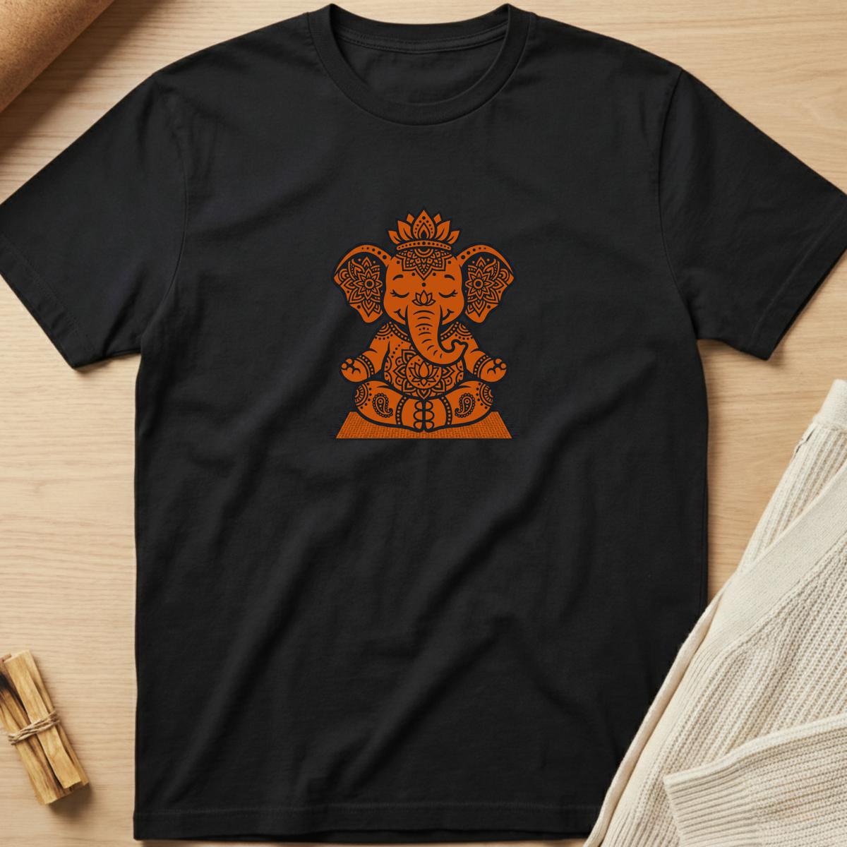 A black short-sleeve crewneck t-shirt featuring a detailed orange Ganesha graphic on the chest, laid flat on a light wood background with a neutral knit fabric and a small bundle of incense in the corners. Minimalist yoga apparel by pemuyo.