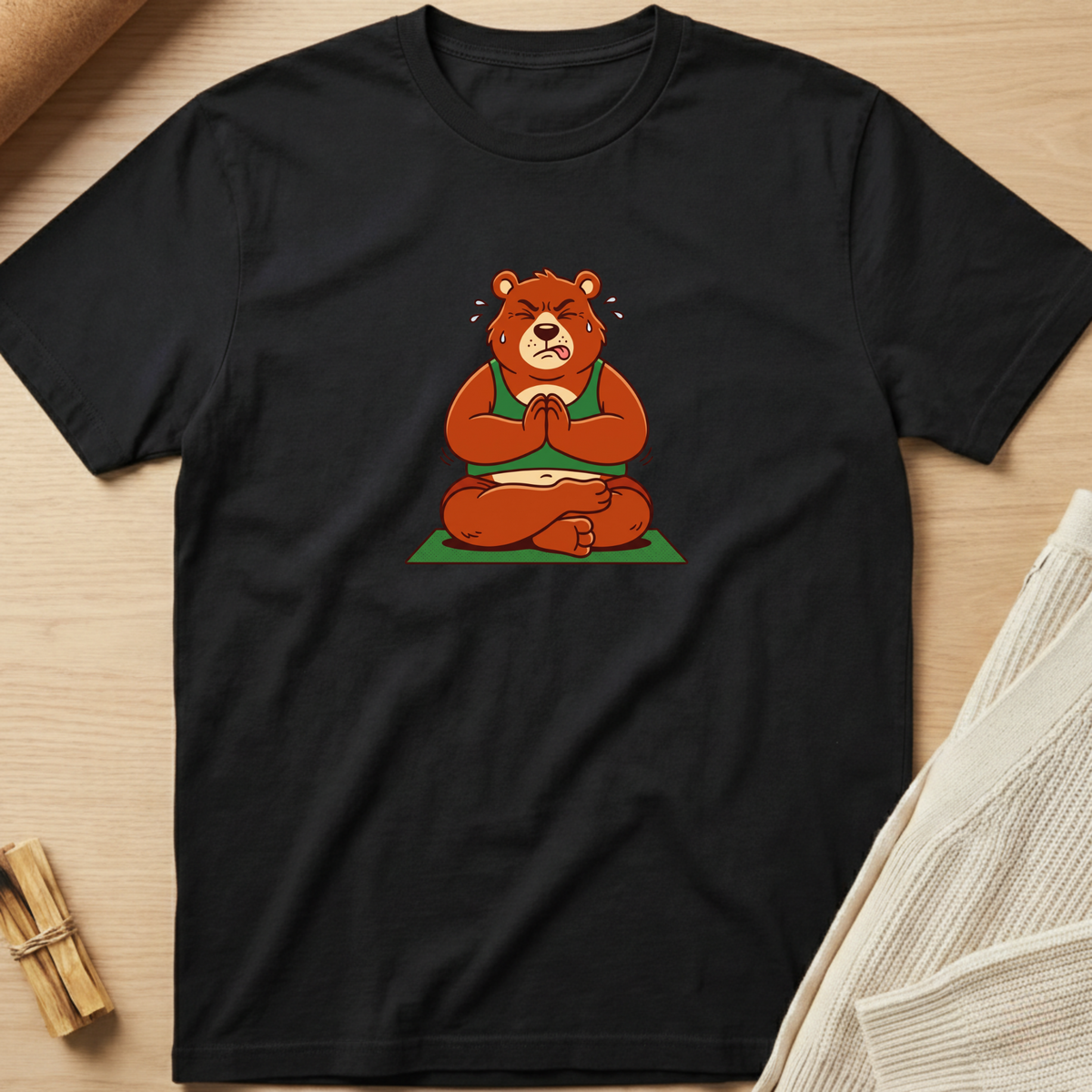A black short-sleeve crewneck t-shirt laid flat on a light wood surface, featuring a cartoon graphic of a brown teddy bear meditating in a green outfit. Minimalist yoga apparel by pemuyo.