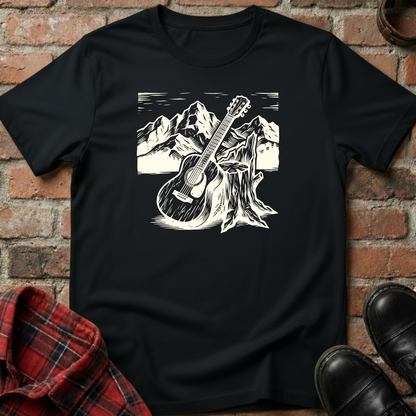 Black t-shirt with a white graphic of a guitar leaning against a tree stump with mountains in the background, displayed on a brick surface. Minimalist music apparel by pemuyo.