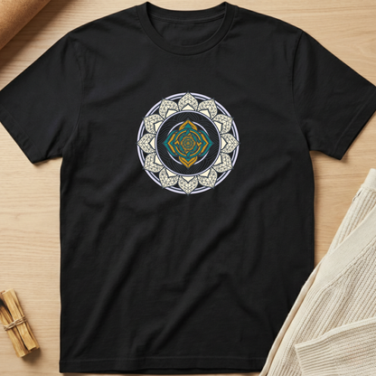 A black short-sleeve crewneck t-shirt laid flat on a light wood surface, featuring a circular mandala-style graphic on the chest with white, gold, and teal details. Minimalist yoga apparel by pemuyo.