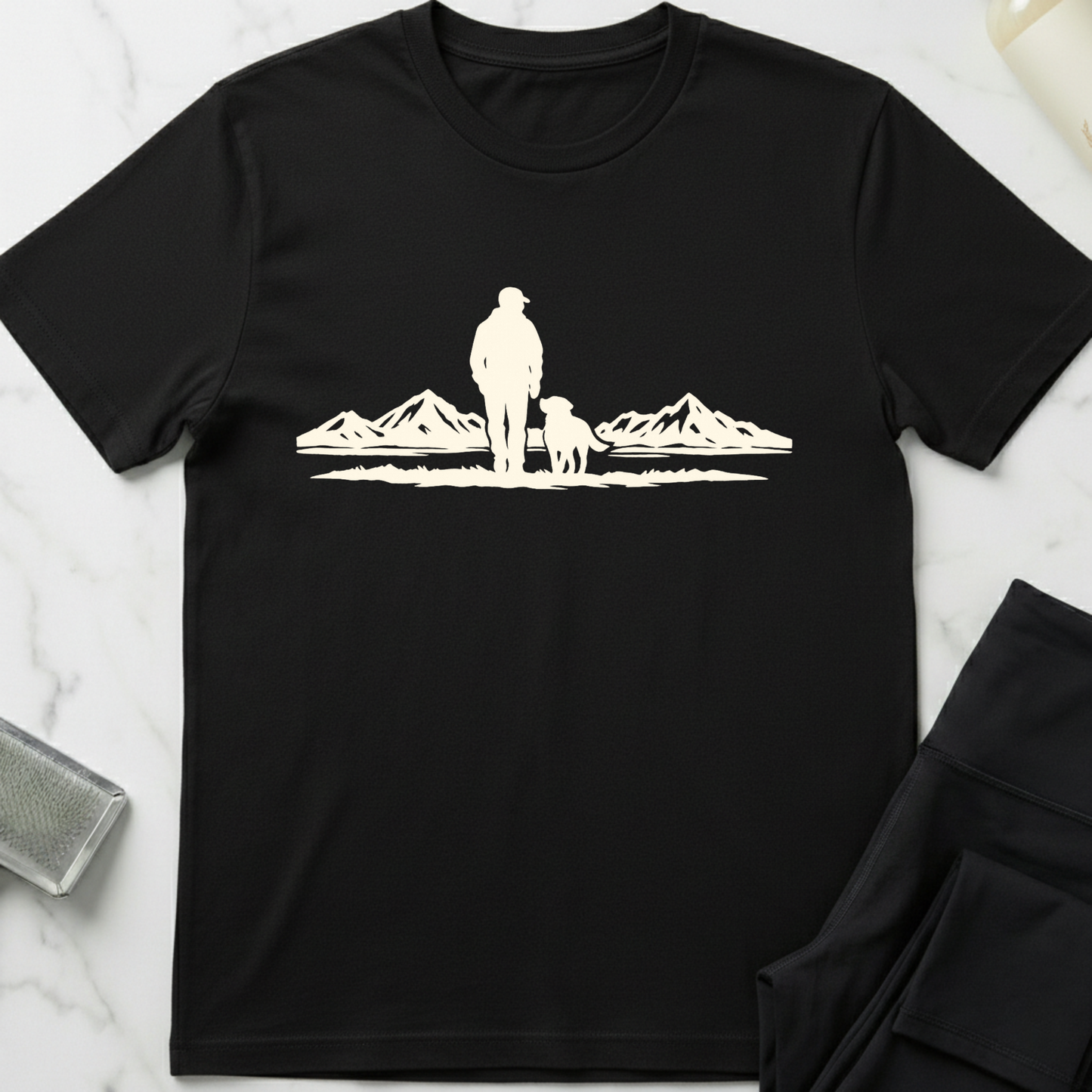 Black t-shirt with white graphic of a person and dog standing in front of mountains on a marble surface. Minimalist dog apparel by pemuyo.