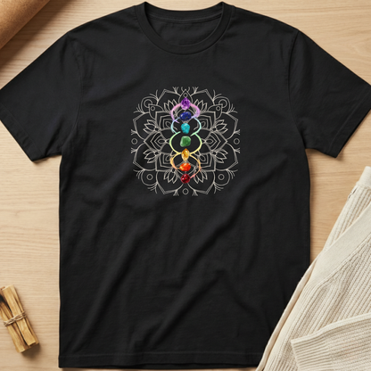 A black short-sleeve t-shirt laid flat on a light wood surface, featuring a white mandala-style chakra graphic with a vertical stack of colorful gem-like symbols in the center. A cream knit fabric and small wooden props are partially visible around the edges. Minimalist yoga apparel by pemuyo.
