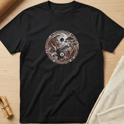 A black short-sleeve t-shirt laid flat on a light wood surface, featuring a circular yin-yang style graphic on the chest with a skull, moon, and dragon design in brown, beige, and white tones. Minimalist yoga apparel by pemuyo.