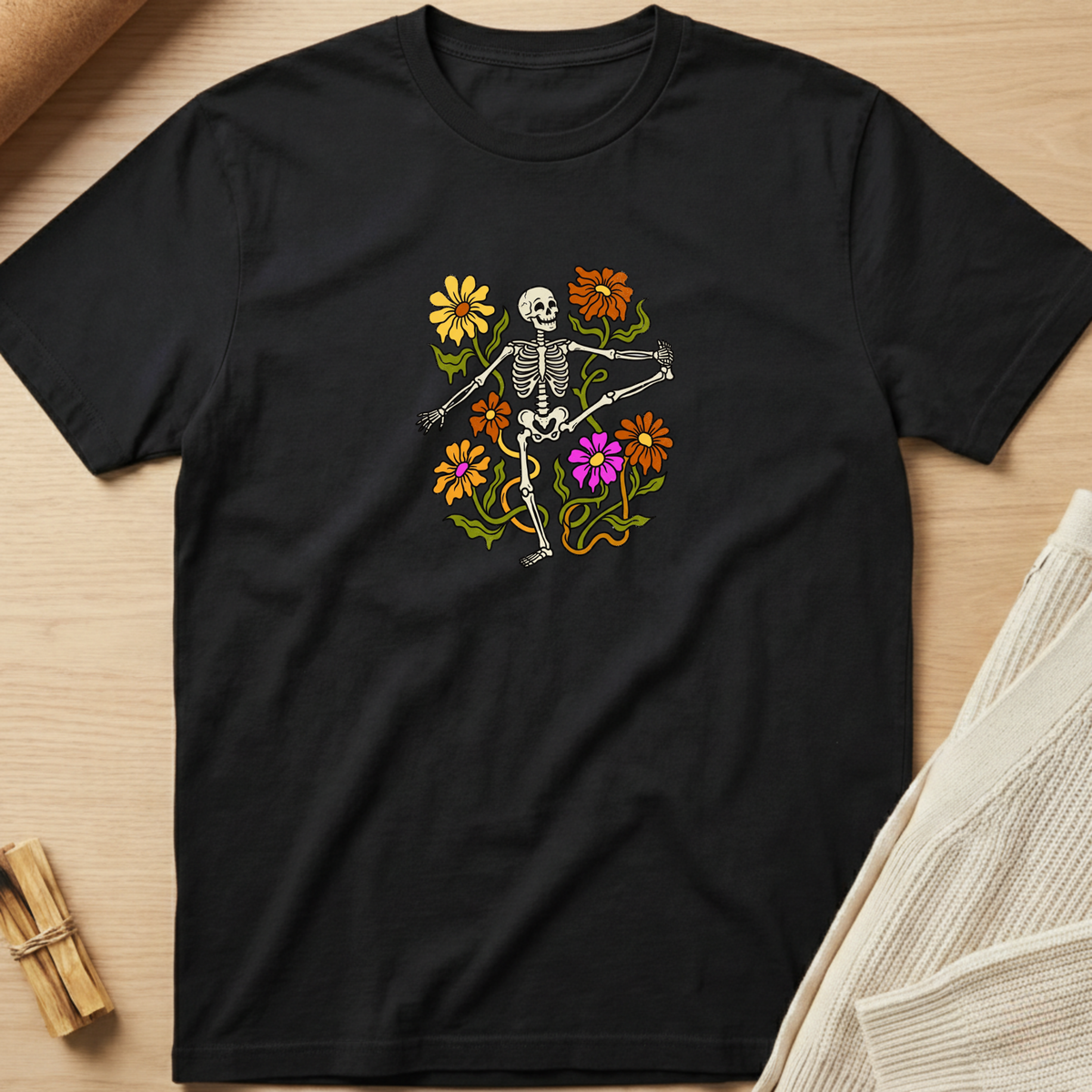 A black short-sleeve crewneck t-shirt laid flat on a light wood surface, featuring a colorful graphic of a dancing skeleton surrounded by yellow, orange, and purple flowers. Minimalist yoga apparel by pemuyo.