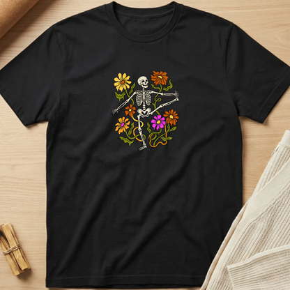 A black short-sleeve crewneck t-shirt laid flat on a light wood surface, featuring a colorful graphic of a dancing skeleton surrounded by yellow, orange, and purple flowers. Minimalist yoga apparel by pemuyo.