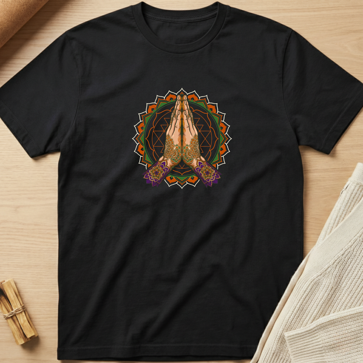 A black short-sleeve t-shirt laid flat on a light wood surface, featuring a colorful graphic of two hands in a prayer pose inside a mandala design. Styled flat-lay props include a rolled beige fabric in the top-left and a folded cream knit blanket in the bottom-right. Minimalist yoga apparel by pemuyo.
