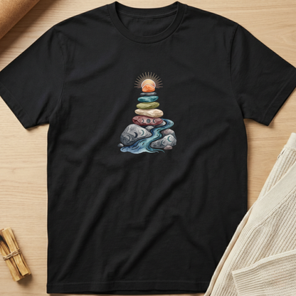 A black short-sleeve crewneck t-shirt laid flat on a light wood background, featuring a central graphic of stacked stones with a sun and flowing blue water. Minimalist yoga apparel by pemuyo.