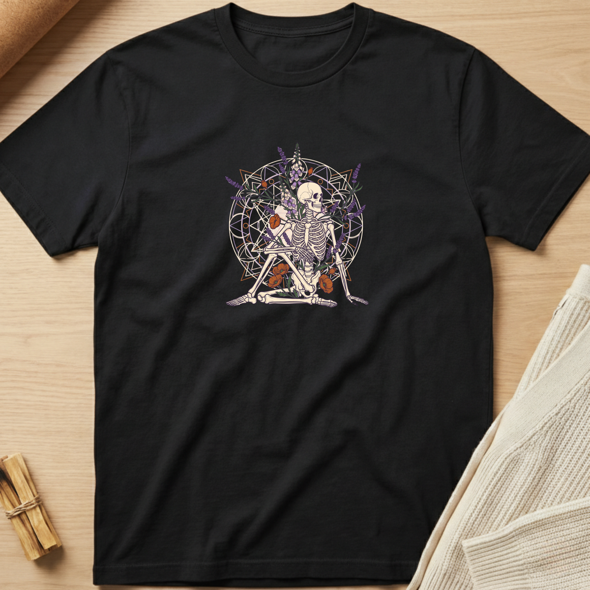 A black short-sleeve t-shirt laid flat on a light wood surface, featuring a central graphic of a seated skeleton surrounded by occult-style symbols and purple lightning bolts. Minimalist yoga apparel by pemuyo.