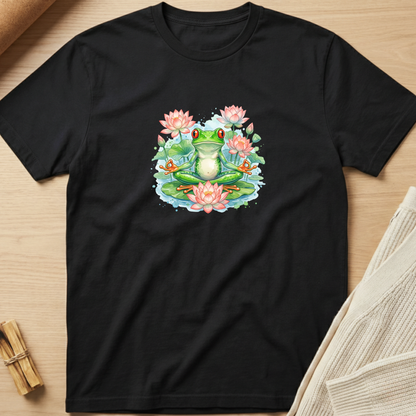 A black short-sleeve t-shirt laid flat on a light wood surface, featuring a colorful graphic of a green frog meditating in a pond with pink water lilies. Minimalist yoga apparel by pemuyo.