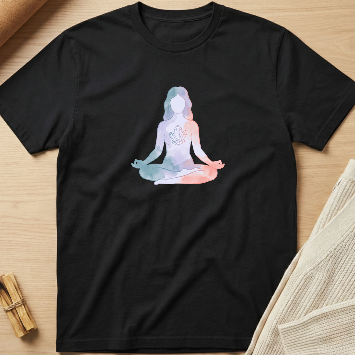 A black short-sleeve crewneck t-shirt laid flat on a light wood surface, featuring a pastel watercolor-style graphic of a meditating figure in shades of blue, pink, and white. The shirt is styled with a beige knit sweater and small wooden props. Minimalist yoga apparel by pemuyo.