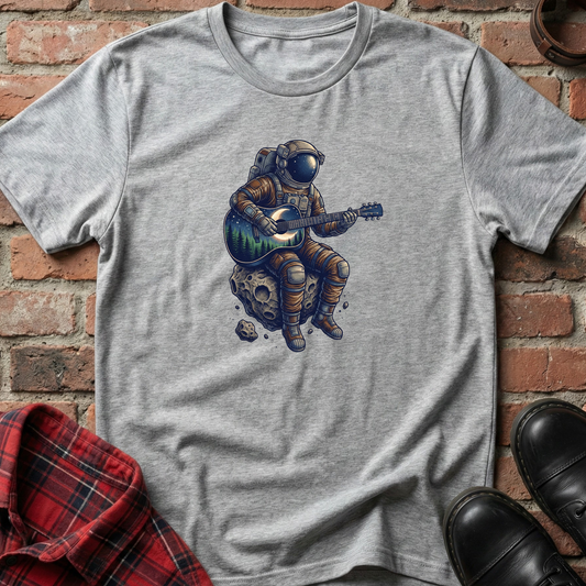 Gray t-shirt with an astronaut playing guitar on a rock design, placed against a brick wall. Minimalist music apparel by pemuyo.