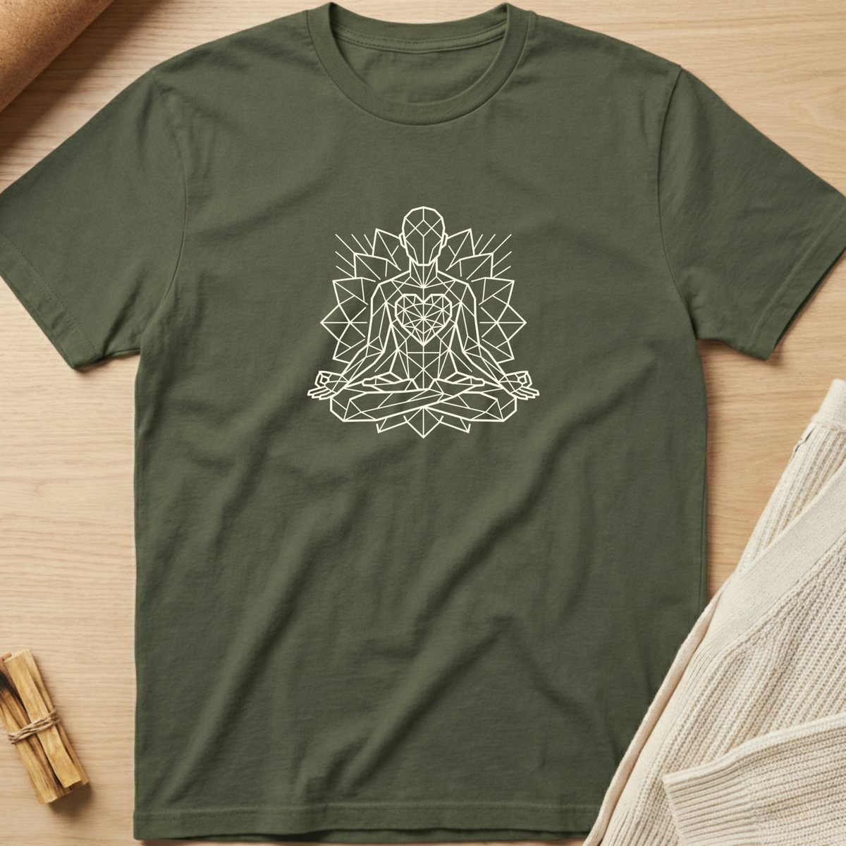 An olive green short-sleeve t-shirt with a white geometric line art graphic of a meditating figure, laid flat on a light wood background with neutral props. Minimalist yoga apparel by pemuyo.