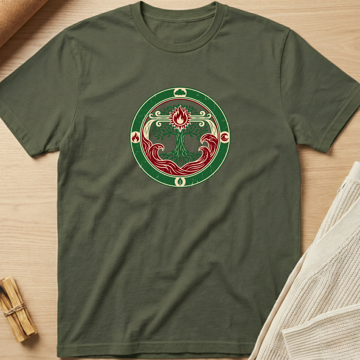 An olive green crewneck t-shirt laid flat on a light wood surface, featuring a circular graphic print in the center with a stylized tree of life in red, green, and cream. Minimalist yoga apparel by pemuyo.