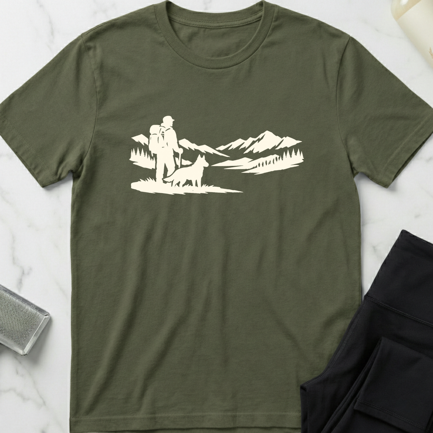 Green t-shirt with white graphic of a person hiking with a dog and mountains on a marble surface. Minimalist dog apparel by pemuyo.