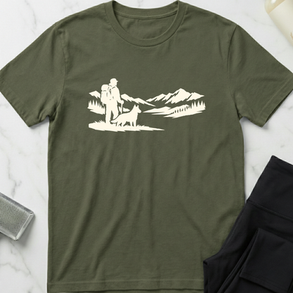 Green t-shirt with white graphic of a person hiking with a dog and mountains on a marble surface. Minimalist dog apparel by pemuyo.