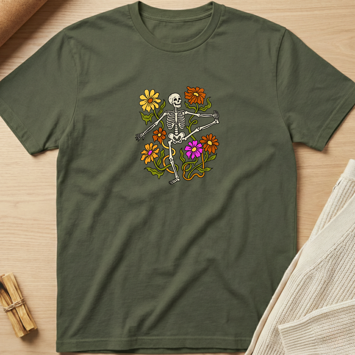 An olive green short-sleeve t-shirt with a colorful graphic of a skeleton dancing among flowers, laid flat on a light wood background. Minimalist yoga apparel by pemuyo.