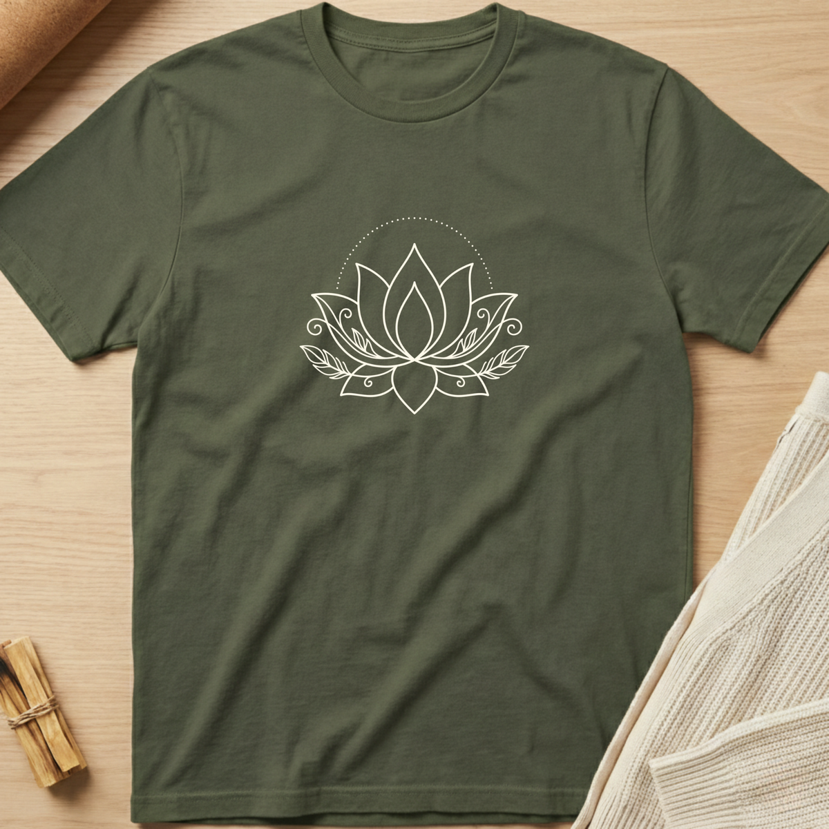 An olive green short-sleeve t-shirt with a white line art lotus flower graphic on the chest, laid flat on a light wood background with styled props. Minimalist yoga apparel by pemuyo.