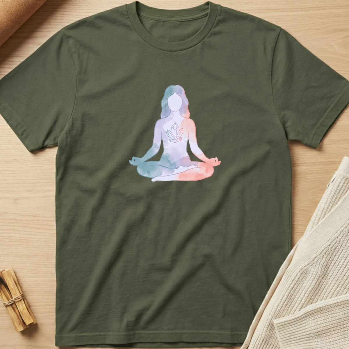 An olive green short-sleeve t-shirt laid flat on a light wood surface, featuring a pastel watercolor-style graphic of a seated meditating figure on the chest. The shirt is styled with neutral props, including a rolled fabric corner, a small bundle of wooden sticks, and a folded cream knit textile. Minimalist yoga apparel by pemuyo.