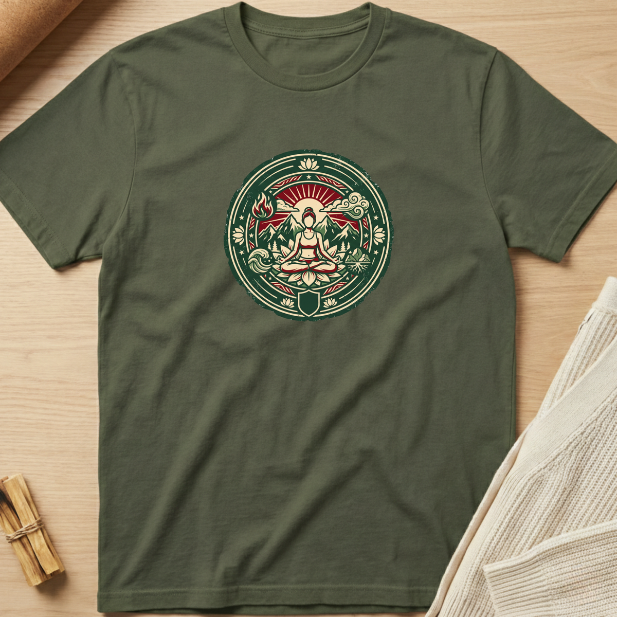 An olive green short-sleeve t-shirt with a circular meditative graphic on the chest, laid flat on a light wood background with styled props. Minimalist yoga apparel by pemuyo.