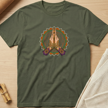 An olive green short-sleeve t-shirt with a central graphic of hands in a prayer pose inside a mandala design, laid flat on a light wood background with styled props. Minimalist yoga apparel by pemuyo.