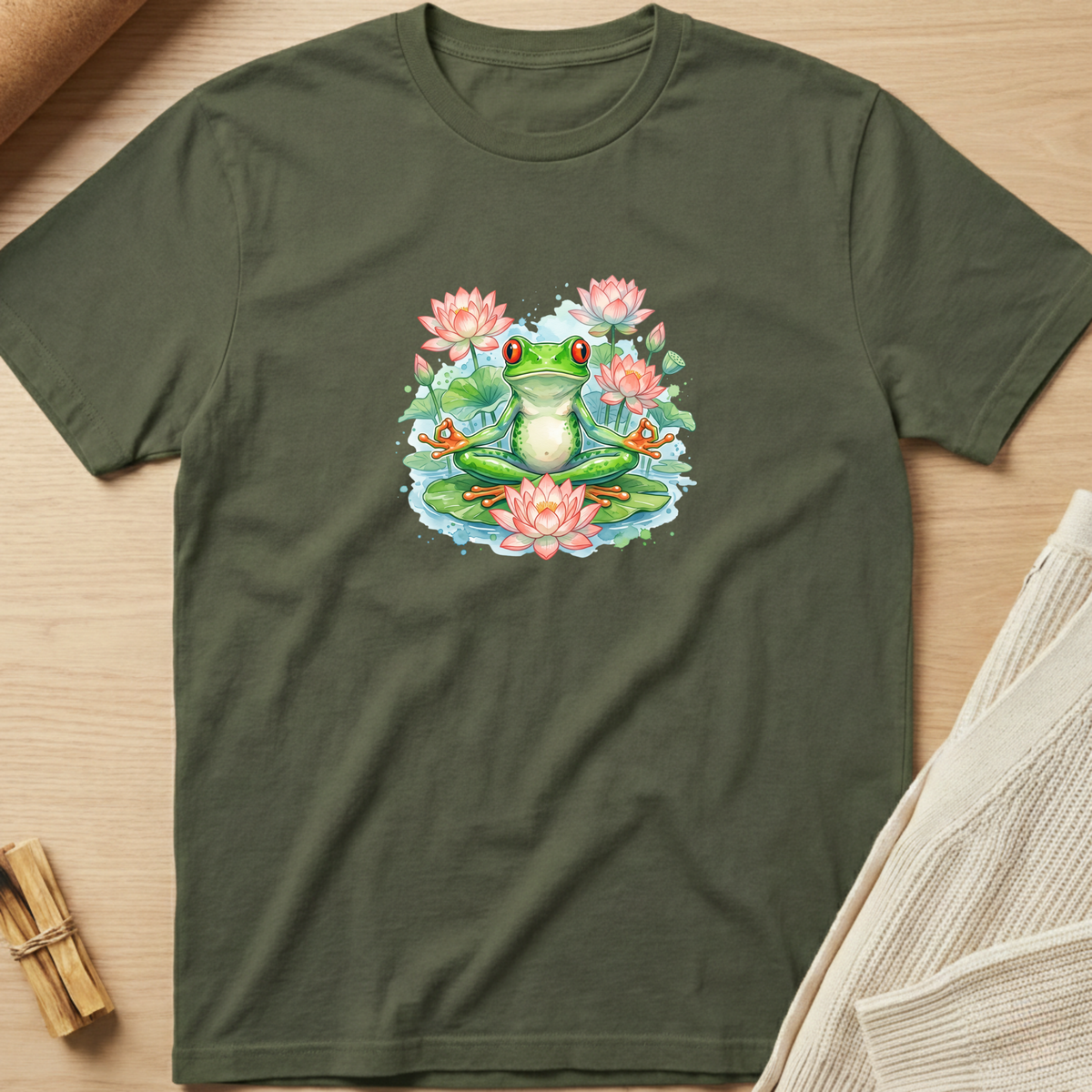 An olive green short-sleeve t-shirt with a colorful graphic of a green frog sitting in a pond with pink lotus flowers, laid flat on a light wood background. Minimalist yoga apparel by pemuyo.