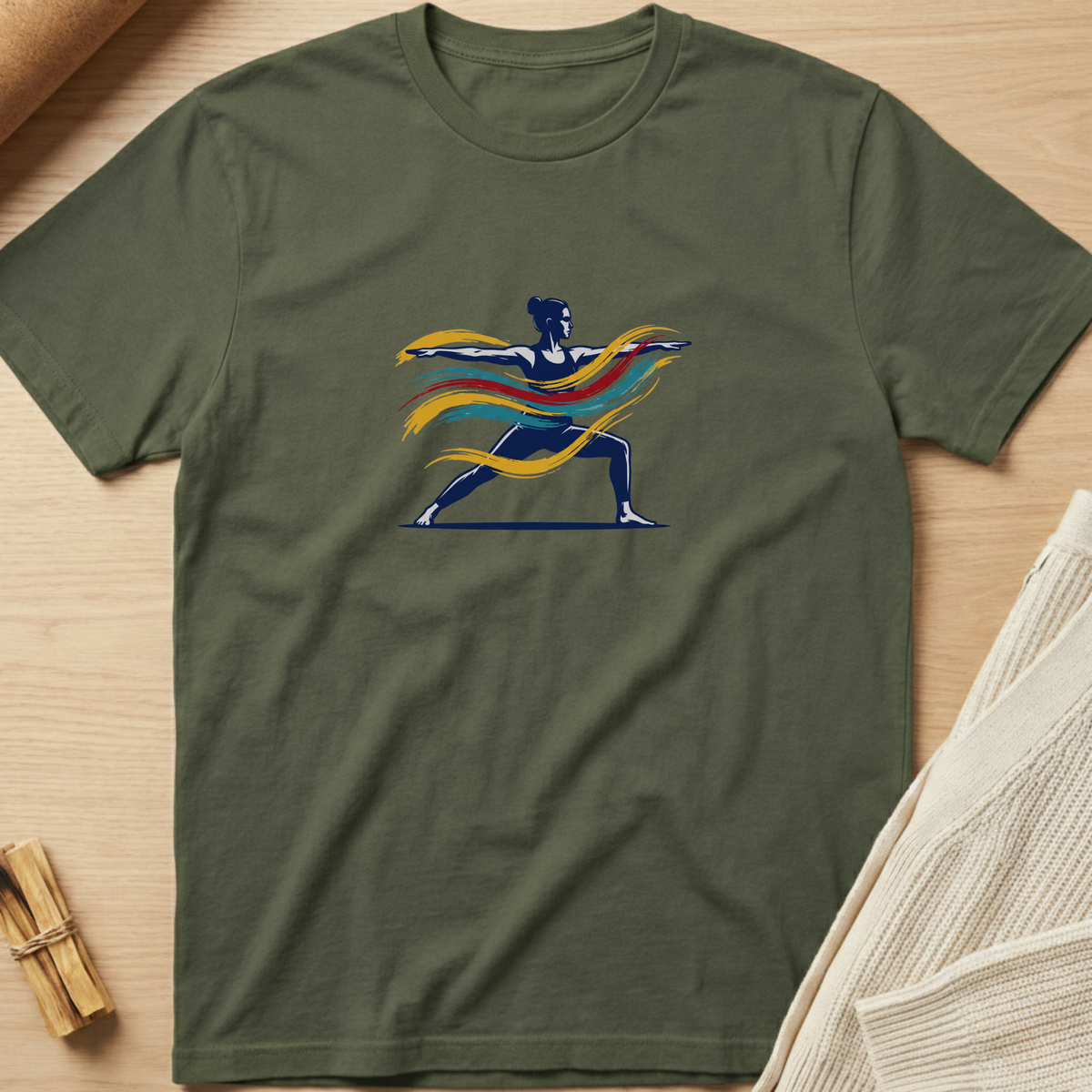 An olive green short-sleeve t-shirt with a graphic of a warrior yoga pose filled with flowing yellow, orange, and red stripes, displayed flat on a light wood background. Minimalist yoga apparel by pemuyo.