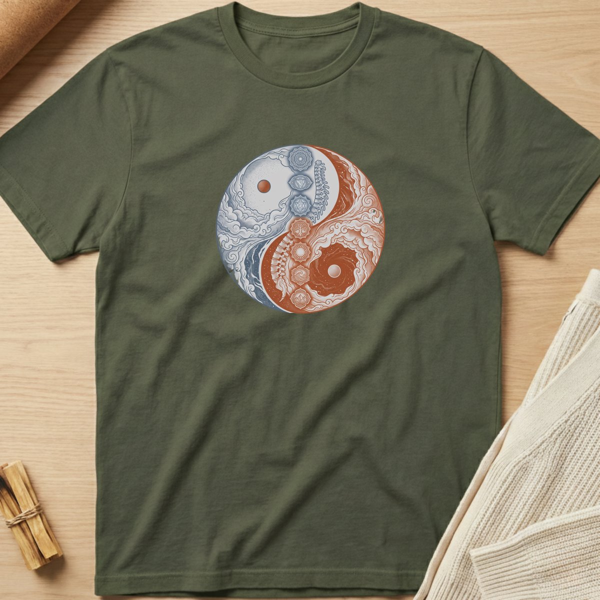 An olive green short-sleeve t-shirt laid flat on a light wood surface, featuring a circular yin-yang style graphic on the chest with blue and orange floral and geometric patterns. Minimalist yoga apparel by pemuyo.