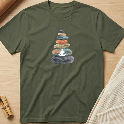 An olive green short-sleeve t-shirt laid flat on a light wood surface, featuring a watercolor graphic of stacked stones with a white meditating figure in the center. Minimalist yoga apparel by pemuyo.
