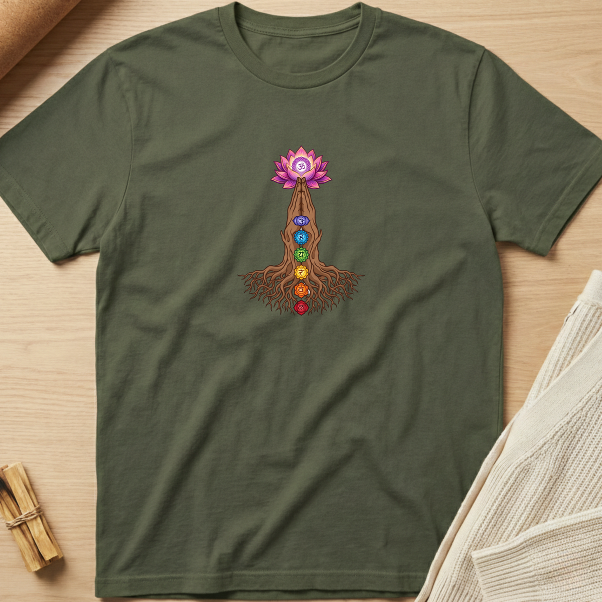 An olive green short-sleeve t-shirt with a colorful chakra and lotus flower graphic on the chest, laid flat on a light wood background with styled props. Minimalist yoga apparel by pemuyo.