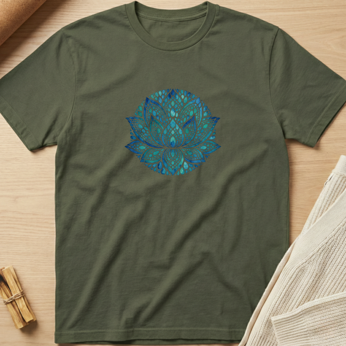 An olive green short-sleeve t-shirt with a blue and teal lotus flower mandala graphic on the chest, laid flat on a light wood background with styled props. Minimalist yoga apparel by pemuyo.