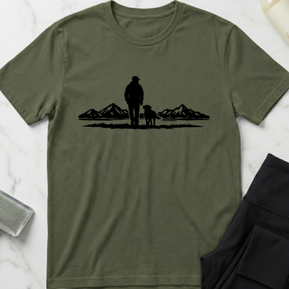 Green t-shirt with a black silhouette of a person and dog walking in a mountainous landscape on a white marble surface. Minimalist dog apparel by pemuyo.