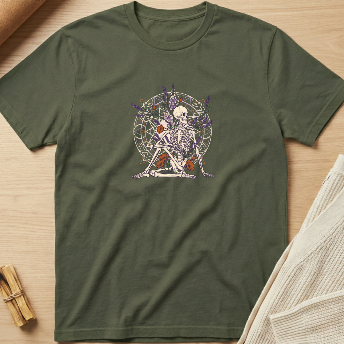 An olive green short-sleeve t-shirt with a central graphic of a skeleton playing a violin in front of a circular occult symbol, laid flat on a light wood background with styled props. Minimalist yoga apparel by pemuyo.