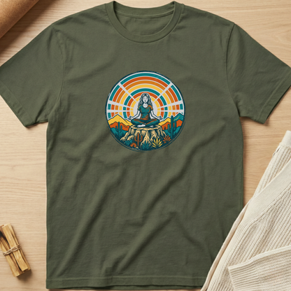 An olive green short-sleeve t-shirt with a colorful circular graphic of a meditating woman on a mountain peak, laid flat on a light wood background with styled props. Minimalist yoga apparel by pemuyo.