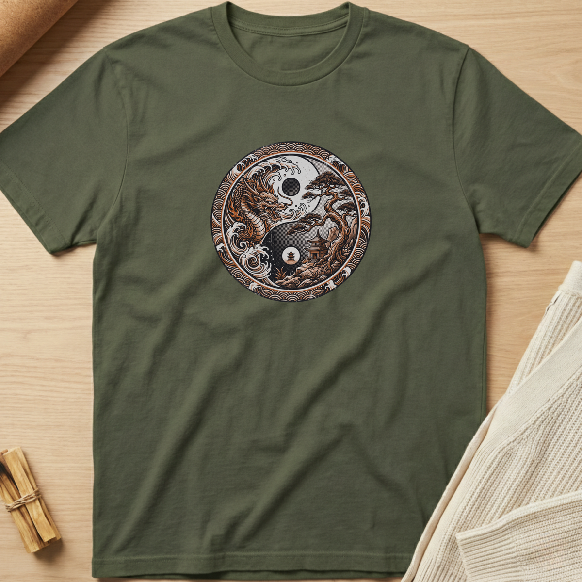 An olive green short-sleeve t-shirt with a circular yin-yang graphic featuring a skull, owl, and star design, laid flat on a light wood background with styled props. Minimalist yoga apparel by pemuyo.