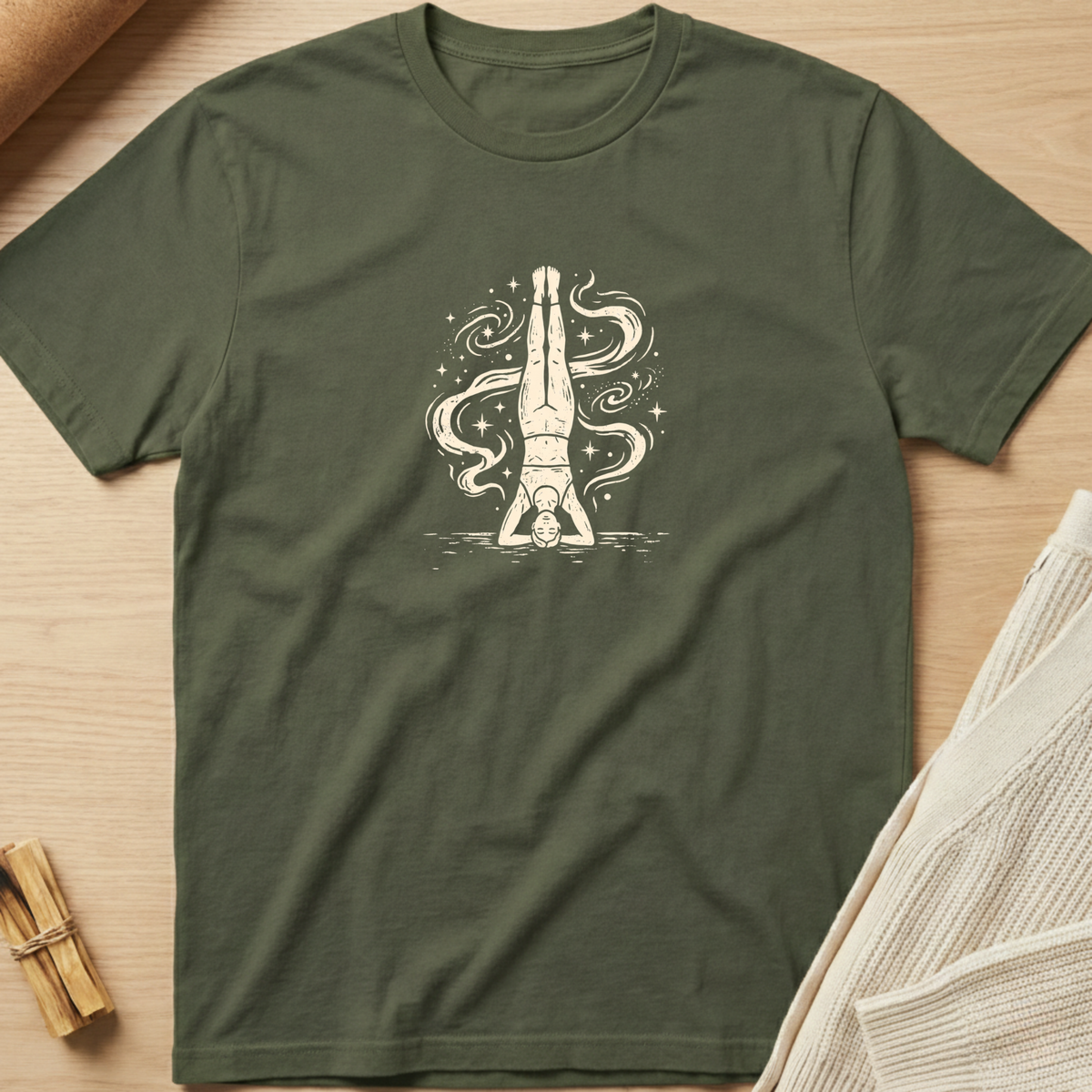 An olive green short-sleeve t-shirt with a cream-colored graphic of a person doing a headstand surrounded by smoke and stars, laid flat on a light wood background with styled props. Minimalist yoga apparel by pemuyo.
