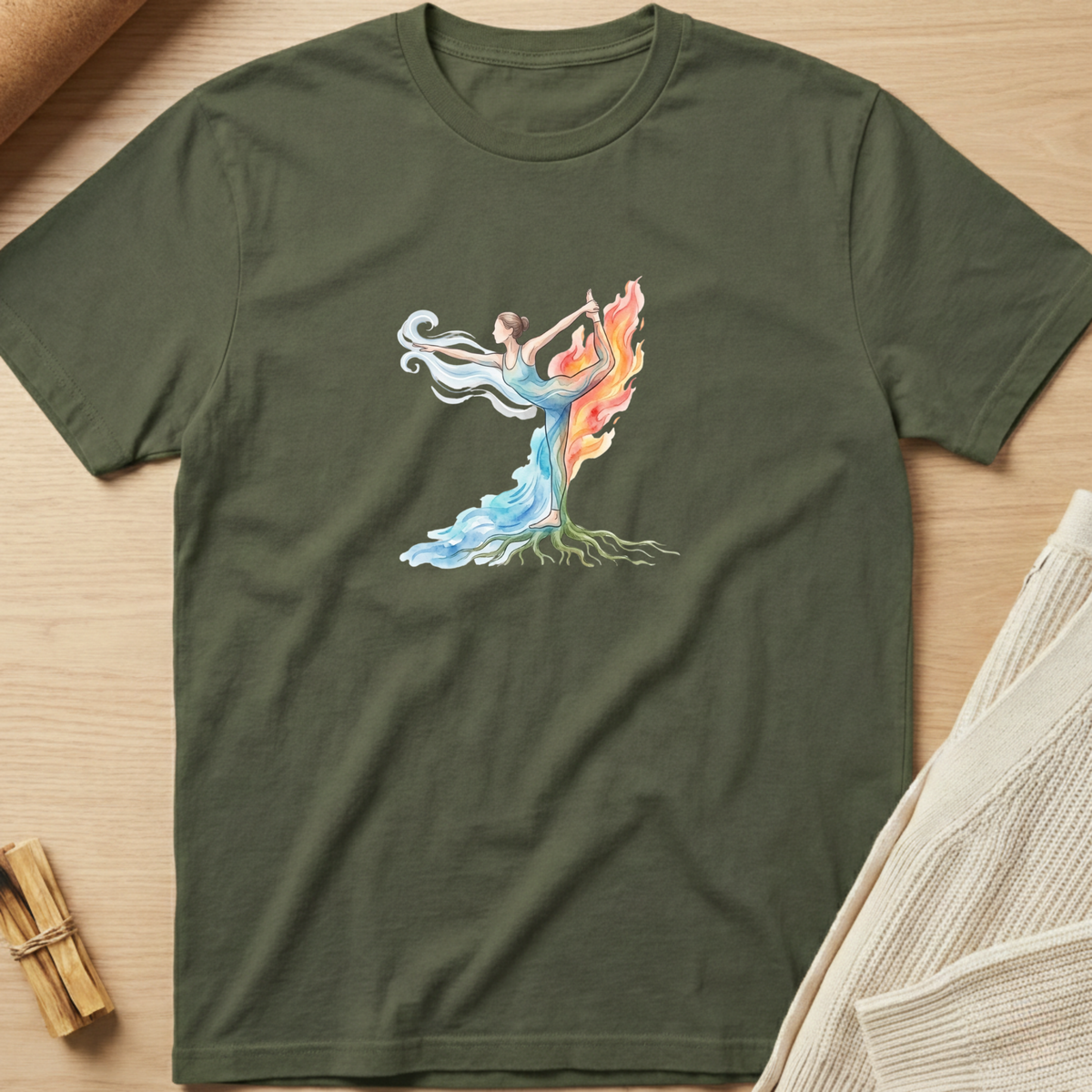An olive green short-sleeve t-shirt with a colorful graphic of a dancing female figure with roots and flowing blue, white, and orange flames, laid flat on a light wood background. Minimalist yoga apparel by pemuyo.