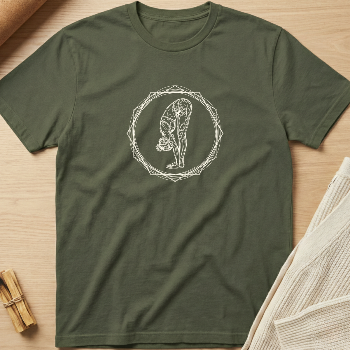 An olive green short-sleeve t-shirt with a white line art graphic of a person in a yoga forward bend pose inside a geometric circle, laid flat on a light wood background with styled props. Minimalist yoga apparel by pemuyo.