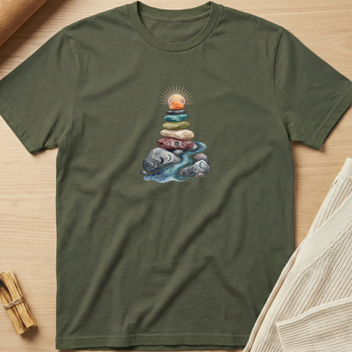 An olive green short-sleeve t-shirt laid flat on a light wood surface, featuring a central graphic of stacked rocks and a flowing blue stream with a small sun motif. Styled flat-lay props include a rolled fabric corner, a small bundle of sticks, and a folded cream knit textile. Minimalist yoga apparel by pemuyo.