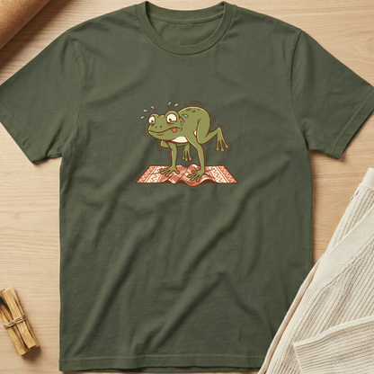 An olive green short-sleeve t-shirt with a cartoon graphic of a sweating green frog standing on a red patterned rug, laid flat on a light wood background with styled props. Minimalist yoga apparel by pemuyo.